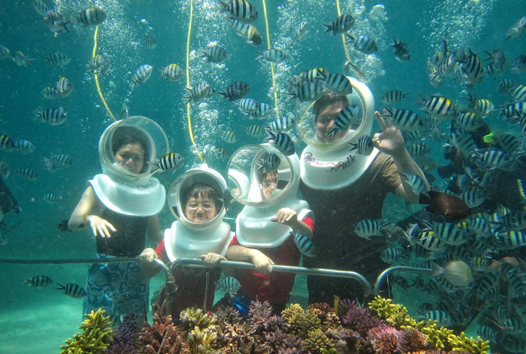 A family with seawalker helmet explore a vibrant coral reef tank, observing the diverse array of sea life swimming beneath seabed of Sanur Bal